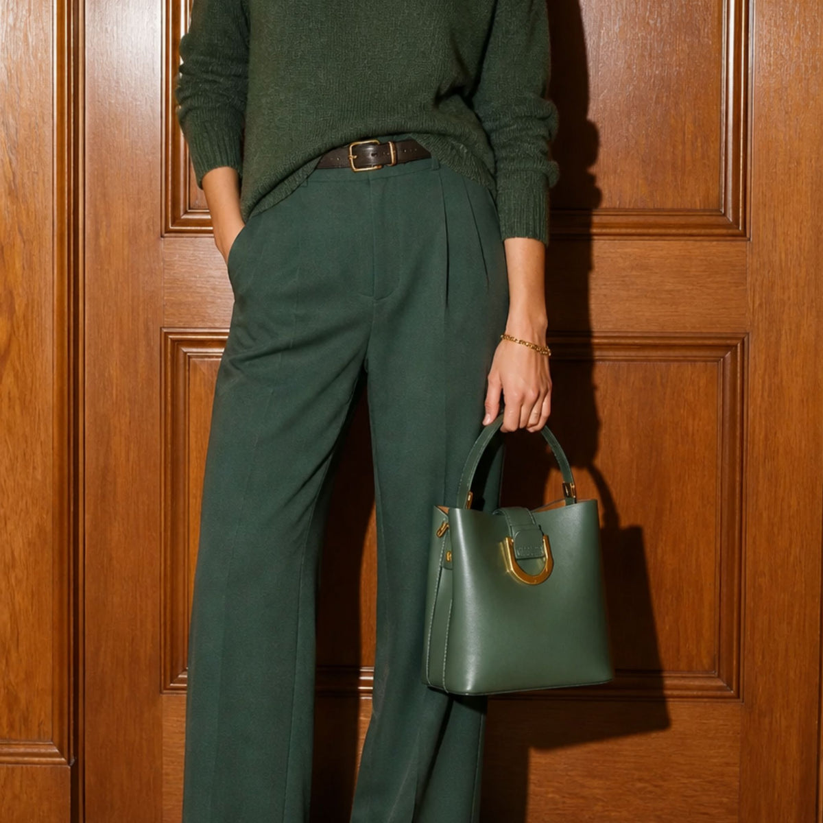 A person wearing a dark green sweater and matching wide-leg trousers stands in front of a wooden door, holding a structured dark green handbag with gold-tone hardware.
