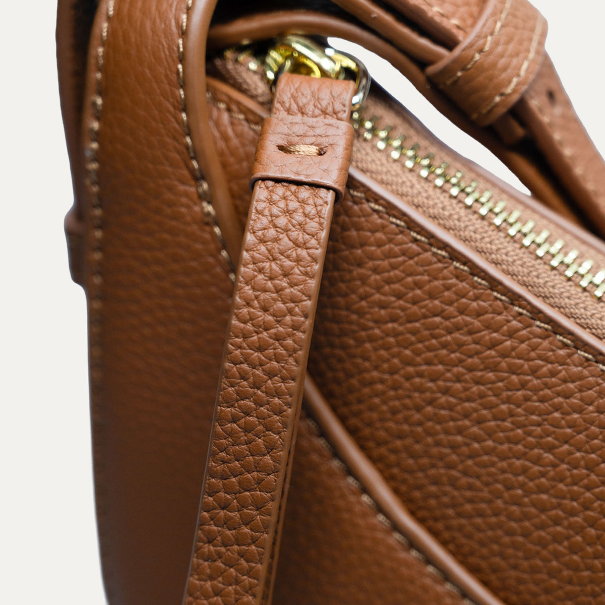 Close-up of a brown pebbled leather handbag showing a strap, zipper, and gold-tone hardware against a light neutral background.