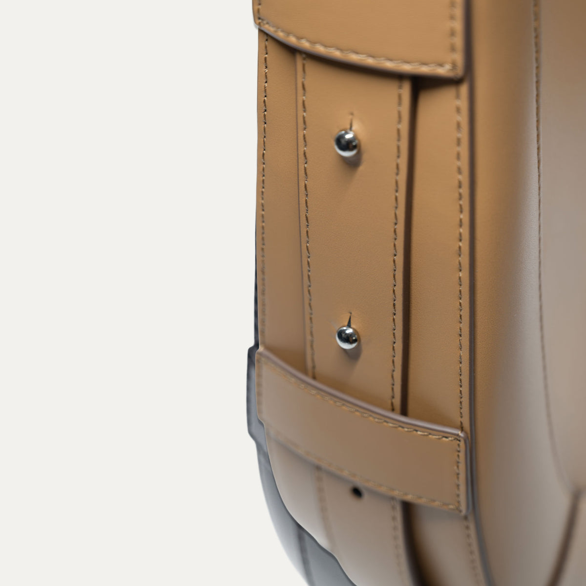 Close-up detail of a tan leather bag strap with silver rivets and contrast stitching on a light gray background.