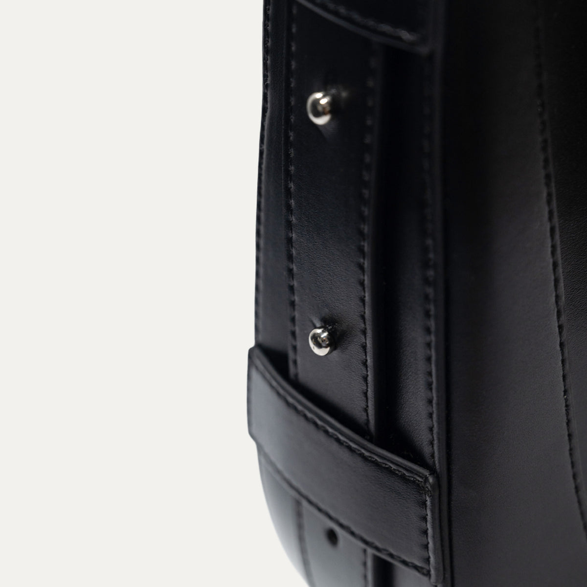 Close-up detail of a black leather bag showing a vertical stitched strap with two silver metal rivets and a matching black leather tab.