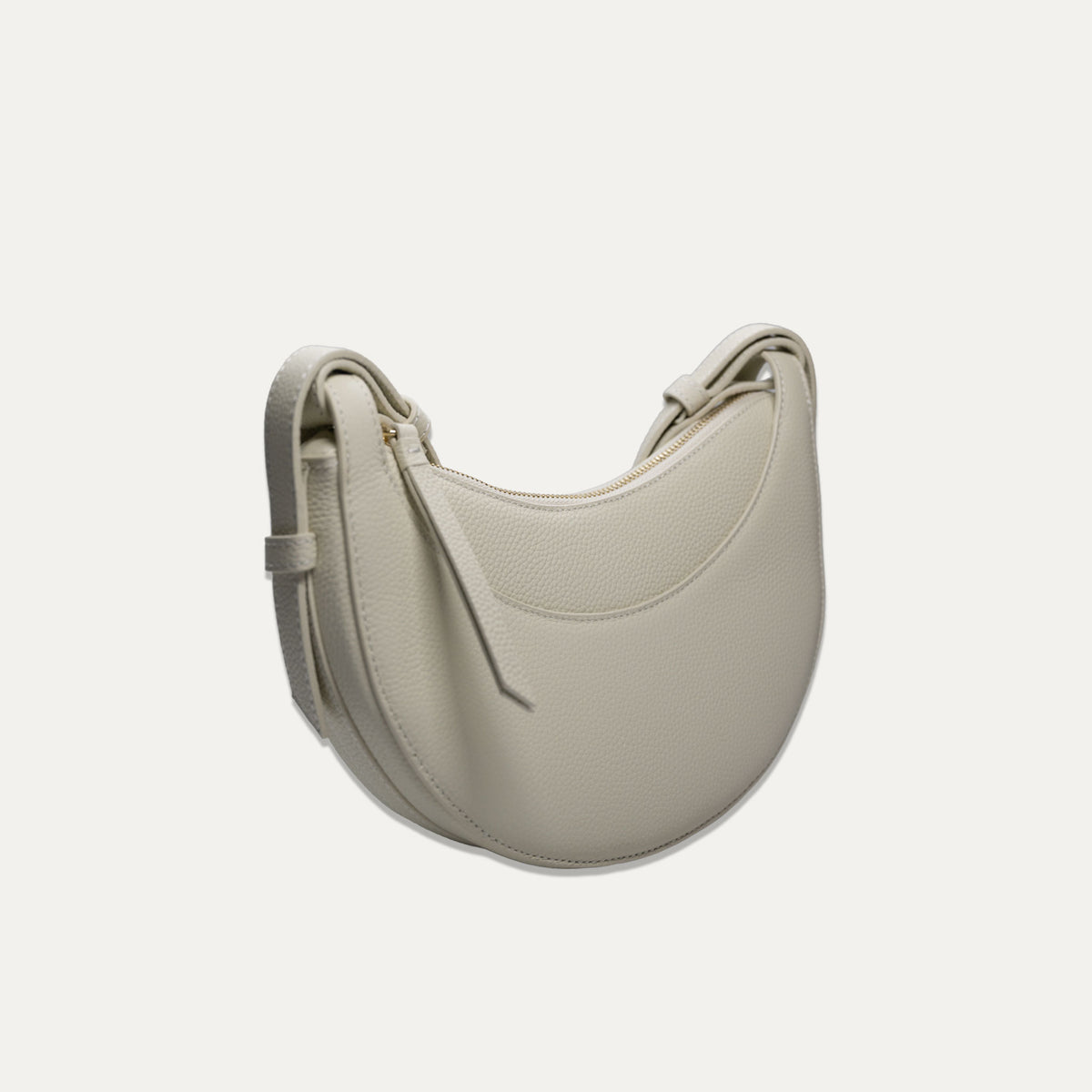 A crescent-shaped pebbled leather shoulder bag in a off-white color, shown against a solid off-white background.