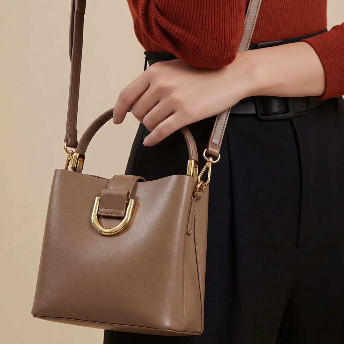 A brown leather bucket handbag with gold hardware, shown being carried by a model wearing a rust-colored top and black trousers.