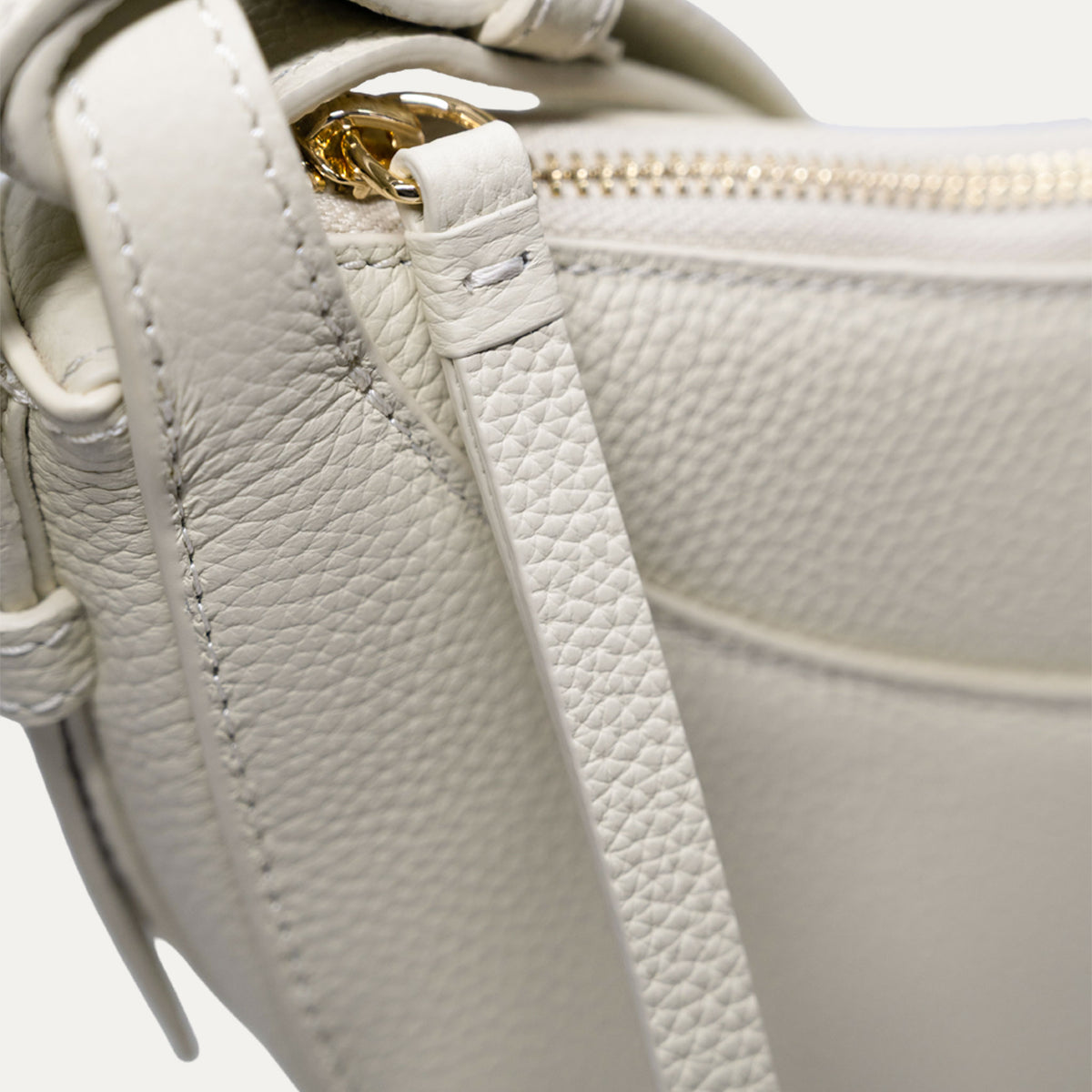 Close-up of a off-white pebbled leather handbag showing the strap attachment with a gold-tone metal ring and a gold zipper.