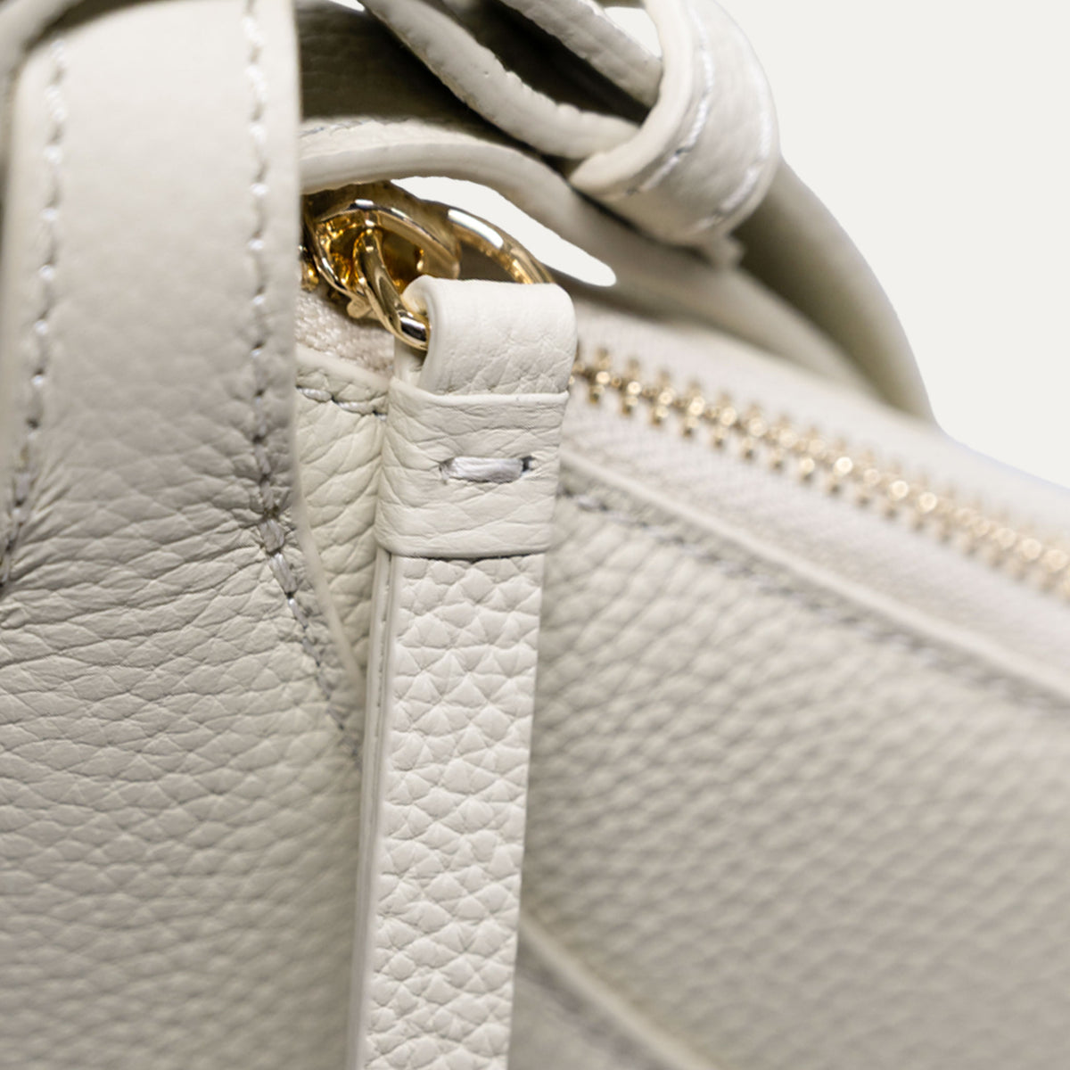 Close-up detail of a off-white pebbled leather handbag showing a strap attachment with gold-tone hardware and a zipper.