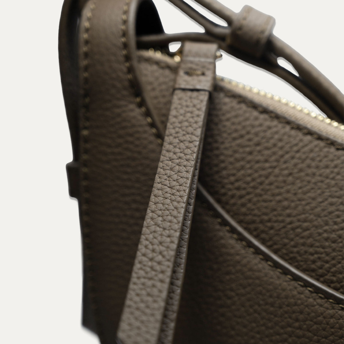 Close-up of a taupe brown pebbled leather handbag showing the strap attachment, zipper, and fine stitching details against a light off-white background.