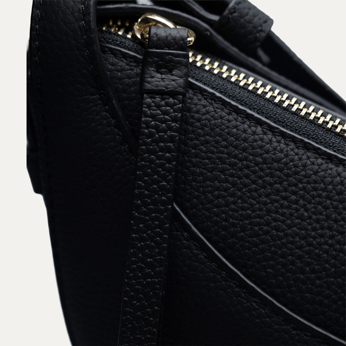Close-up of a black pebbled leather handbag showing a gold-tone zipper and strap attachment on a light gray background.