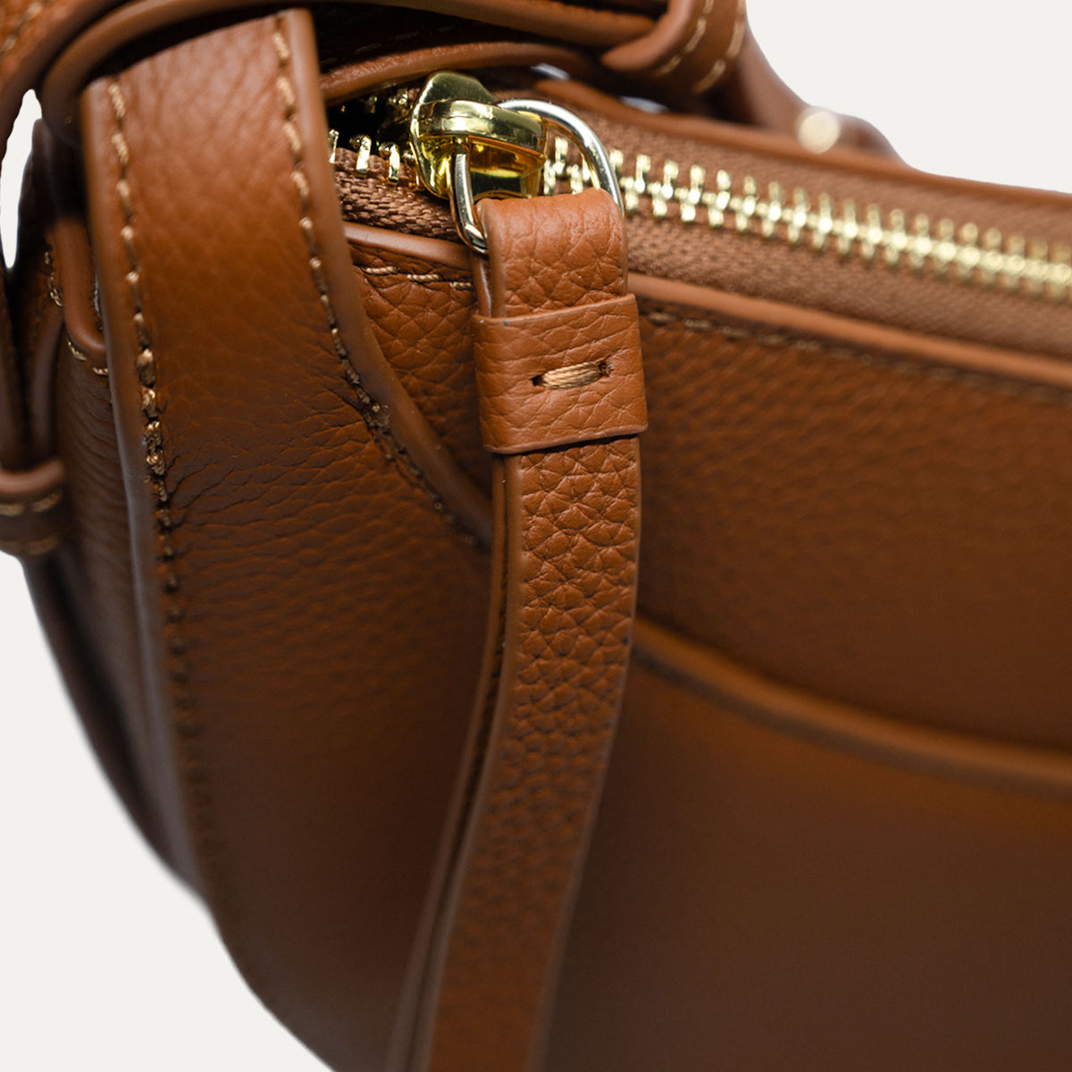 Close-up of a brown pebbled leather handbag showing a gold-tone zipper, metal ring, and matching leather strap detail against a light neutral background.