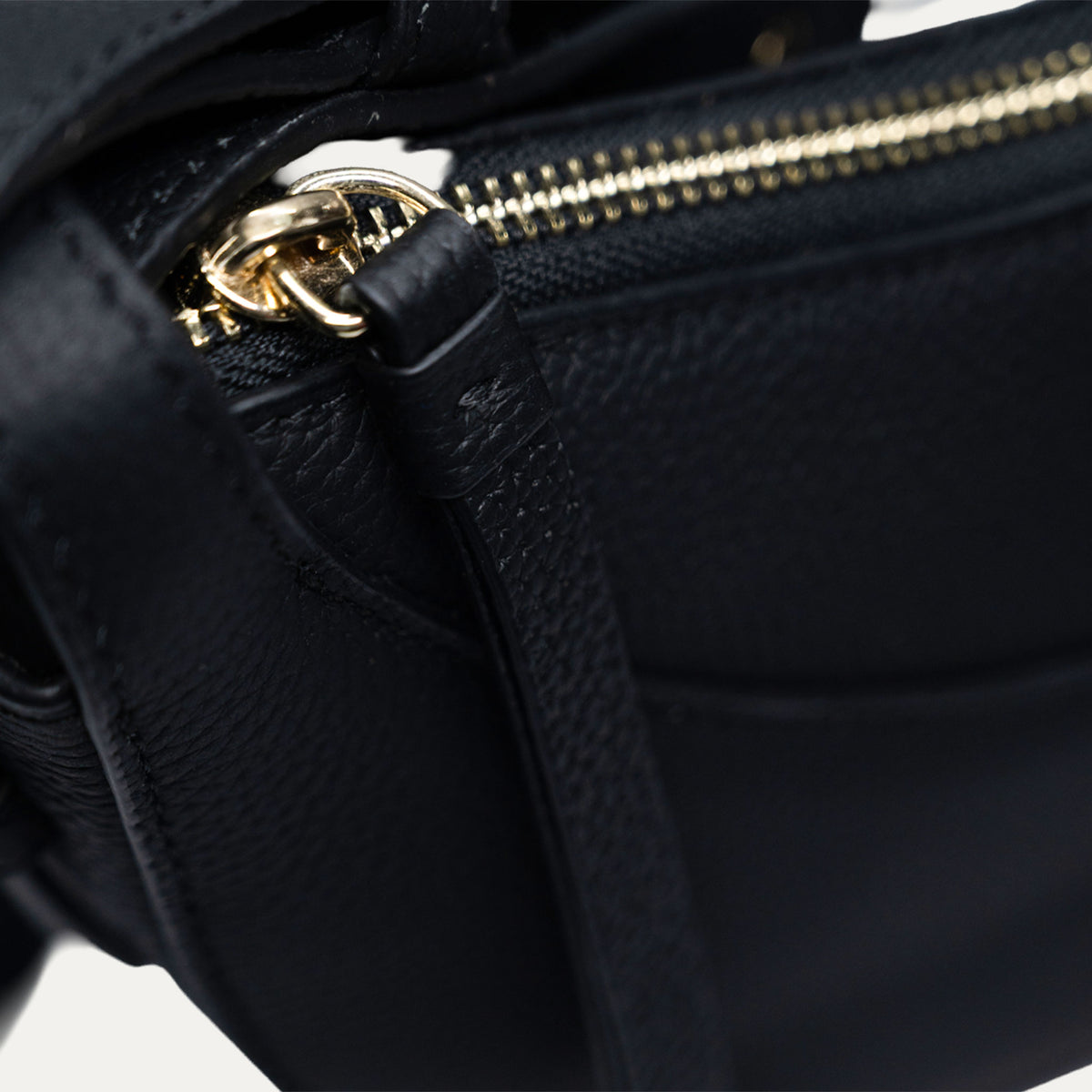 Close-up of a black textured leather bag with a gold-tone zipper and ring pull, showing the strap attachment.