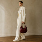 A model stands in a studio wearing a cream-colored high-neck blazer and wide-leg trousers, holding a dark burgundy leather crescent handbag. The background is a textured beige wall with a wooden floor.