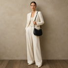 A woman stands against a textured beige wall wearing a cream-colored tailored blazer and wide-leg trousers, holding a black crescent-shaped shoulder bag.